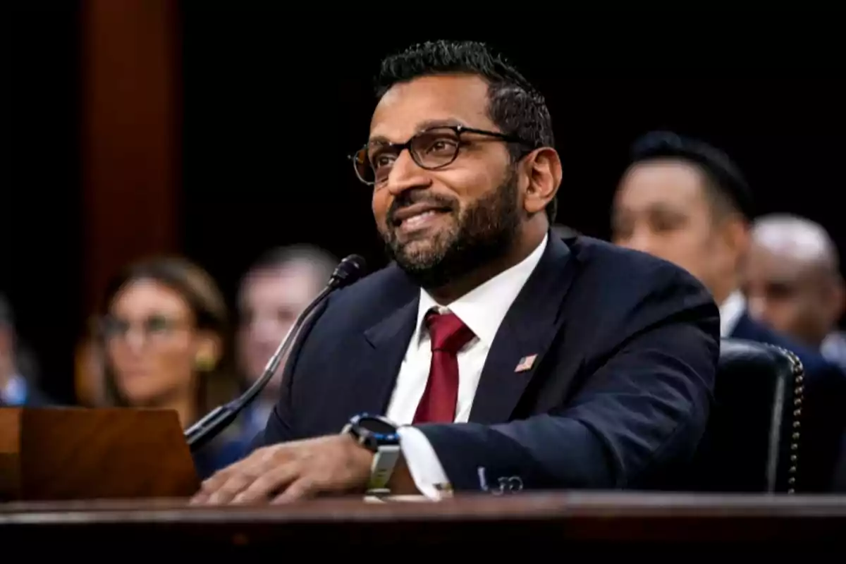 A man in a dark suit and red tie speaks into a microphone during a hearing, with blurred people in the background.