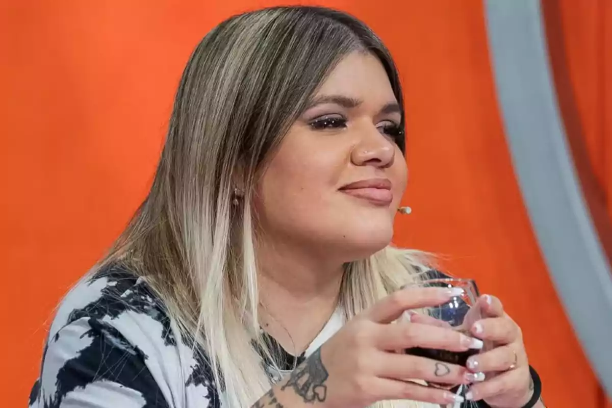 A person with long blonde hair holds a glass while sitting in front of an orange background.