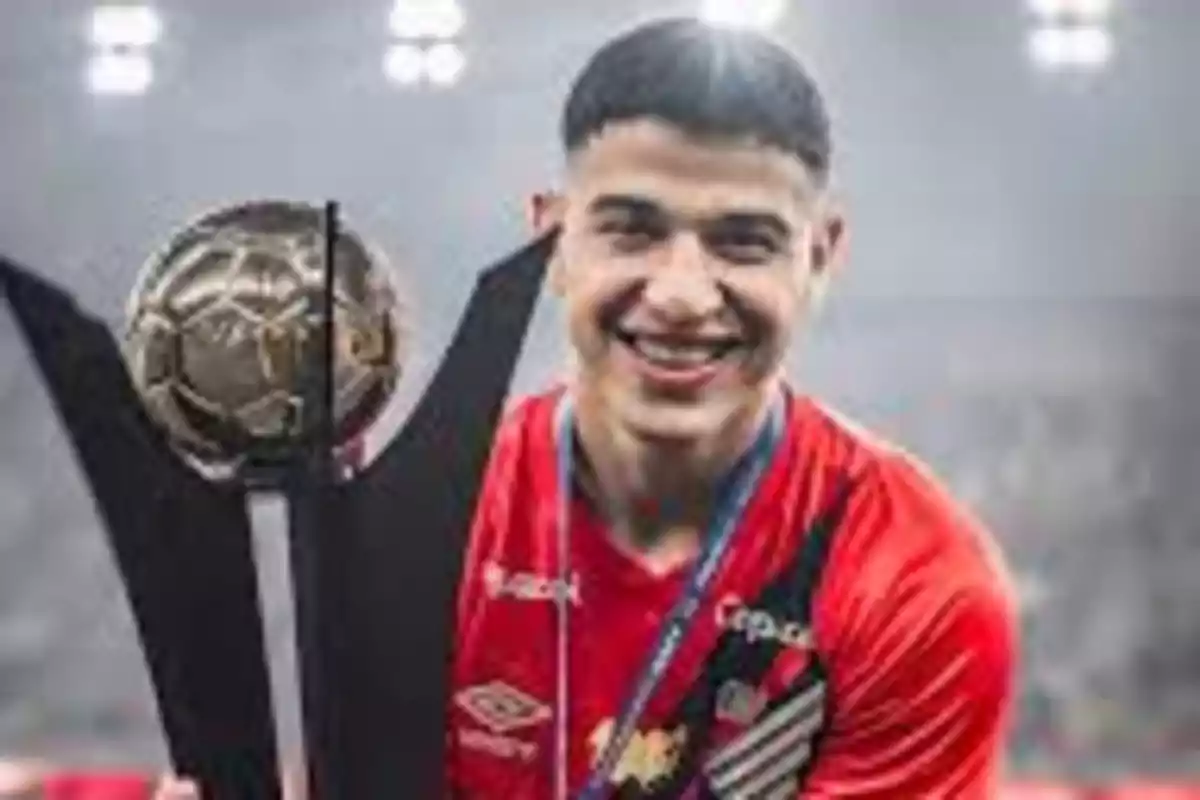 Un jugador de fútbol sonríe mientras sostiene un trofeo en un estadio iluminado.