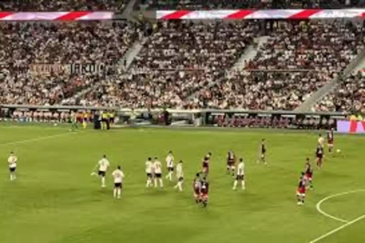 Jugadores de fútbol en el campo durante un partido con un estadio lleno de espectadores.