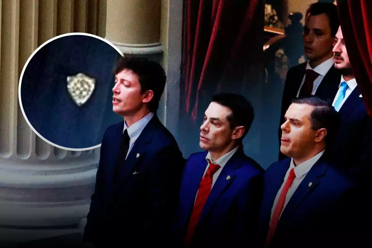 Un grupo de hombres vestidos con trajes oscuros y corbatas de colores variados están de pie frente a unas columnas y cortinas rojas; uno de ellos tiene un pin en la solapa que se muestra en un recuadro ampliado.