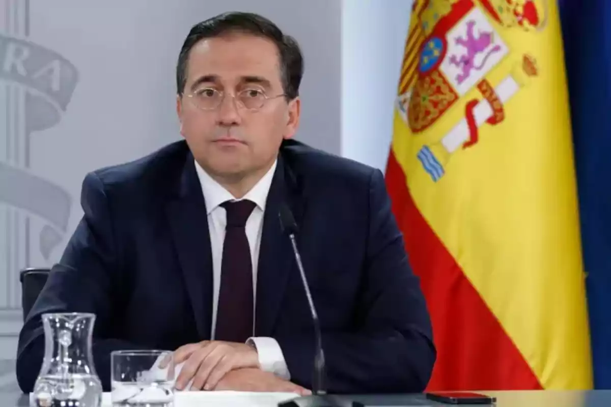 Un hombre con gafas y traje oscuro está sentado frente a un micrófono con una bandera de España en el fondo.