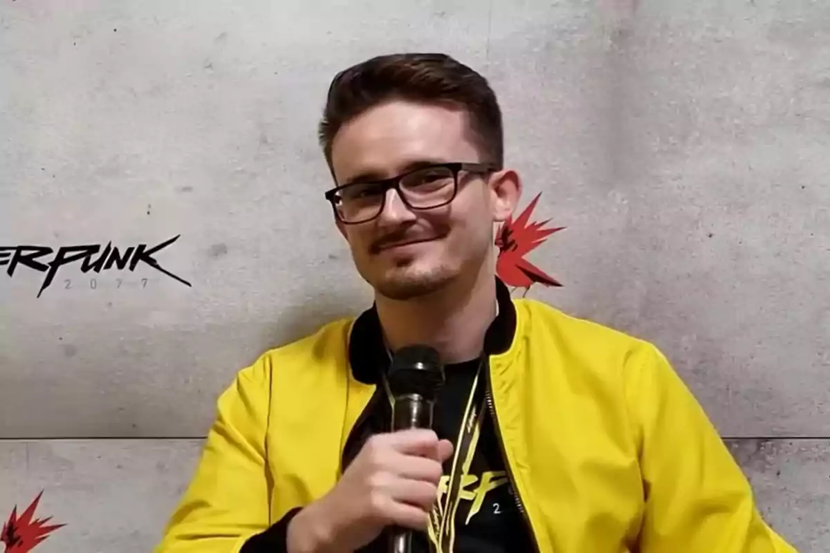 Un hombre con gafas y chaqueta amarilla sostiene un micrófono frente a un fondo con el logo de Cyberpunk 2077.