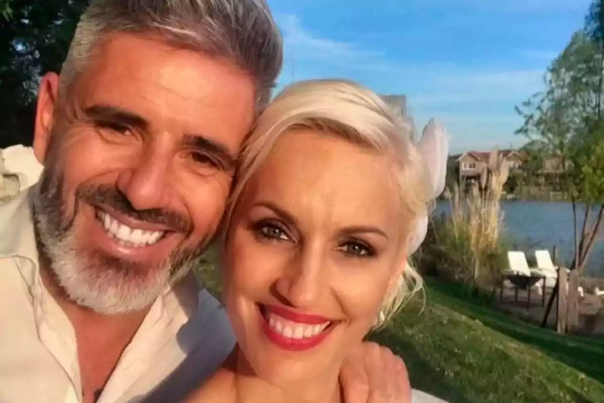 Una pareja sonriente se toma una selfie al aire libre con un paisaje de fondo que incluye césped, árboles y un cuerpo de agua.