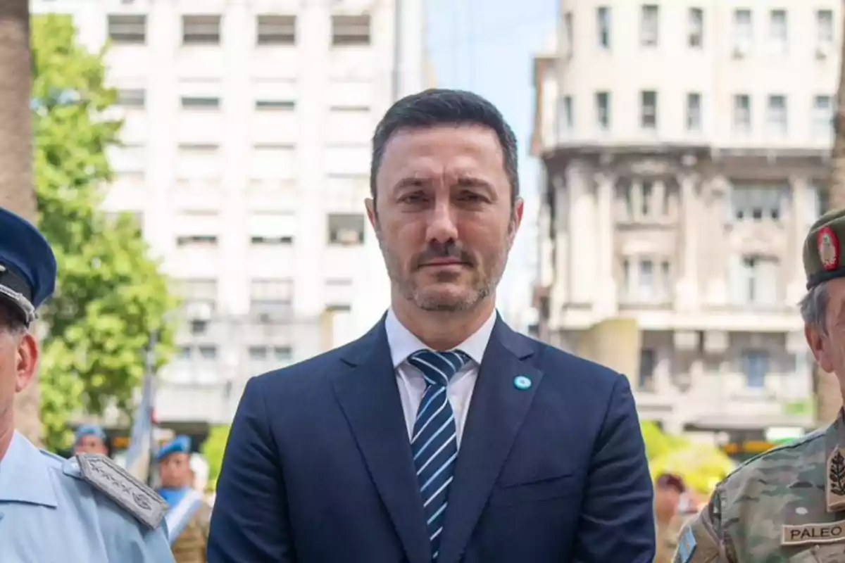 Un hombre de traje y corbata de pie entre dos personas con uniformes militares en un entorno urbano.