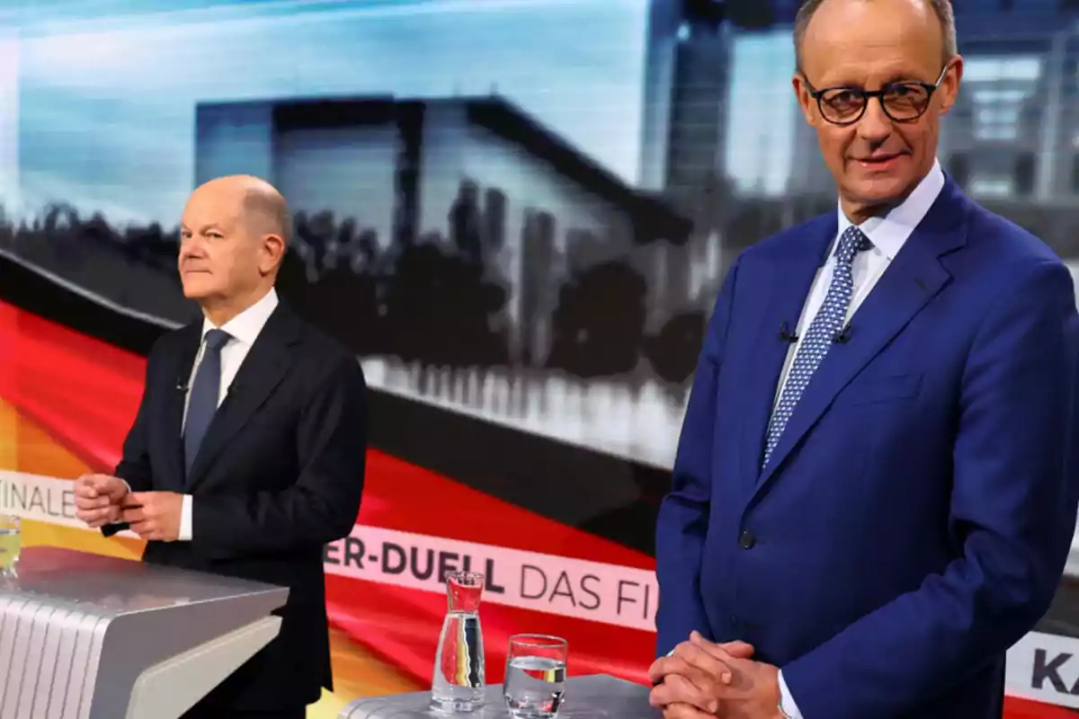 Dos hombres de pie en un escenario de debate con fondo de bandera alemana y edificios desenfocados.