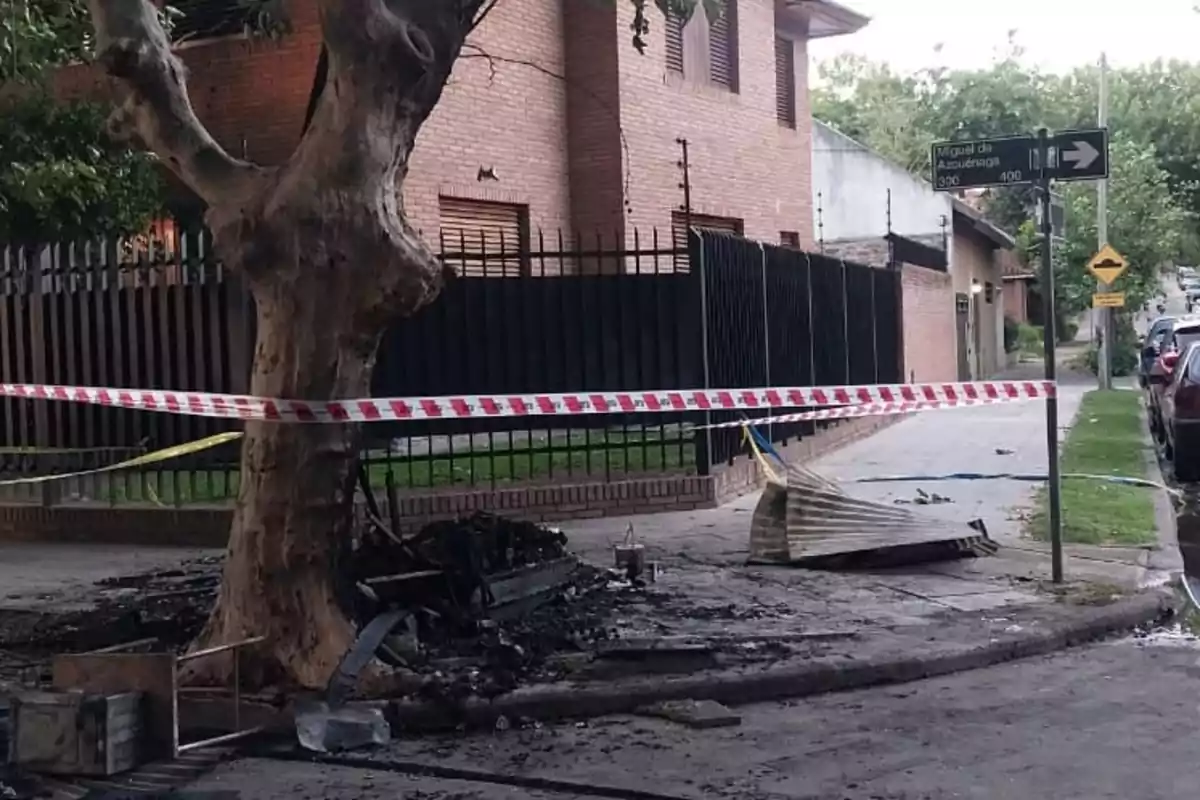 Una escena muestra una acera con restos quemados y escombros junto a un árbol, rodeada por una cinta de seguridad roja y blanca, con una casa de ladrillo y una señal de tráfico en el fondo.