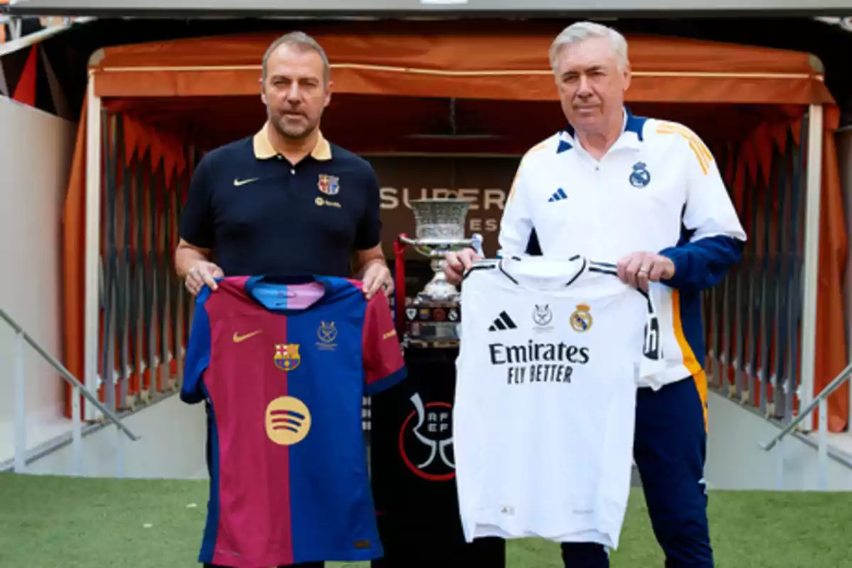 Dos hombres sostienen camisetas de fútbol de los equipos FC Barcelona y Real Madrid frente a un trofeo en un estadio.