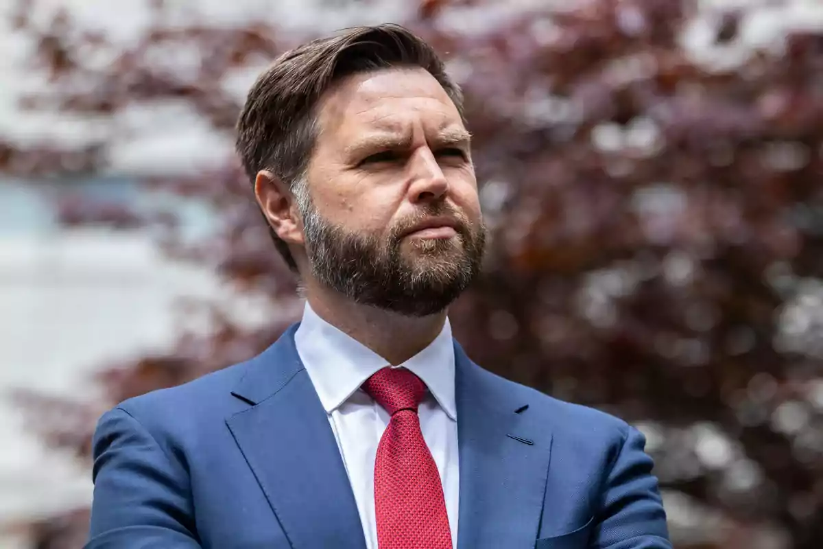 Un hombre con barba y traje azul mira hacia adelante con expresión seria, con un fondo desenfocado de hojas marrones.