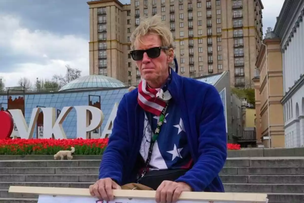 Un hombre con gafas de sol y bufanda con diseño de bandera estadounidense está sentado frente a un edificio alto y un letrero que dice "I ❤️ UKRAINE".