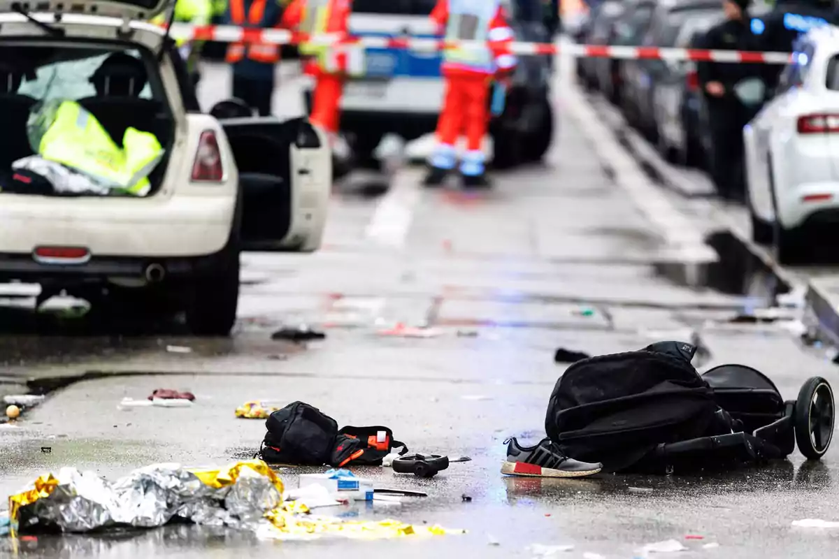 Una escena de accidente en la calle con un coche blanco, objetos personales esparcidos y personal de emergencia al fondo.