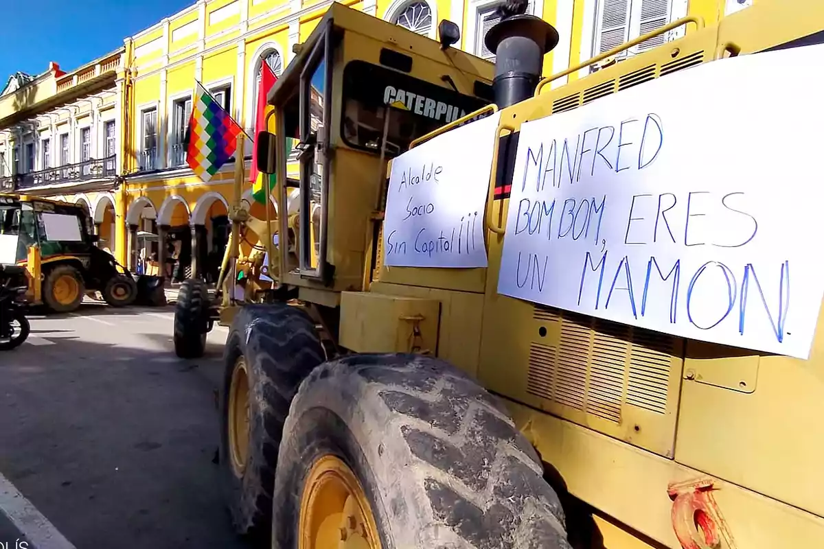 Una protesta con maquinaria pesada en una calle, con carteles escritos a mano y una bandera multicolor.