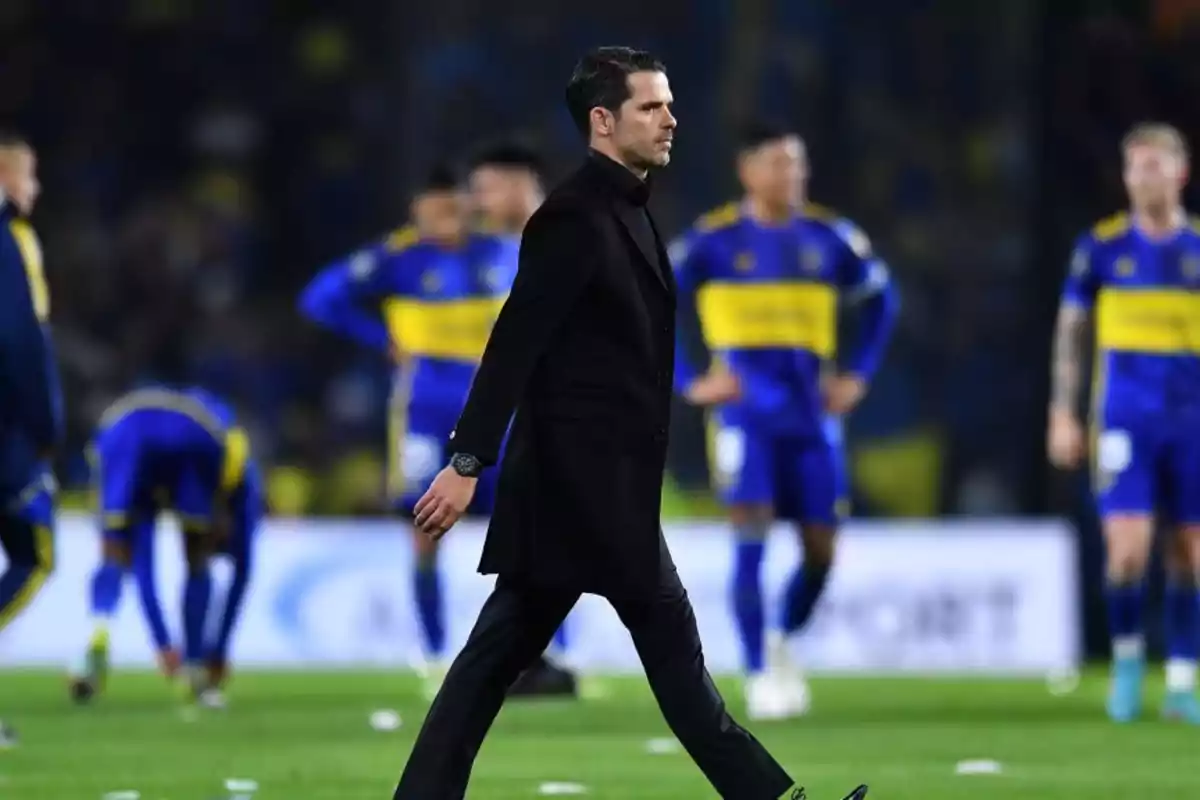 Un hombre vestido de negro camina por el campo de fútbol mientras jugadores con uniformes azules y amarillos están al fondo.