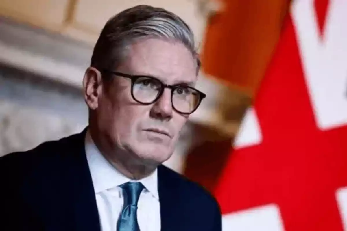 Hombre con gafas y traje oscuro frente a una bandera roja y blanca.