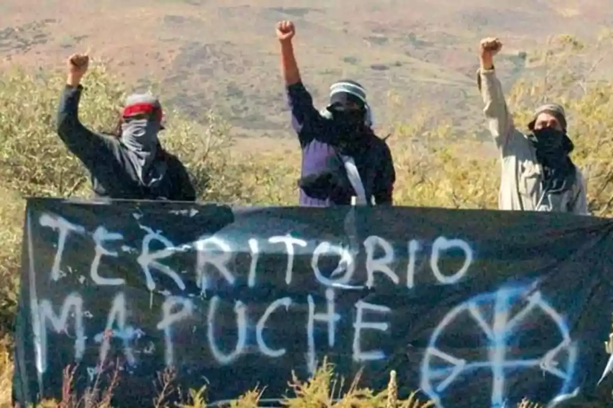 Tres personas con el rostro cubierto levantan el puño detrás de una pancarta negra que dice "Territorio Mapuche" en un paisaje natural.