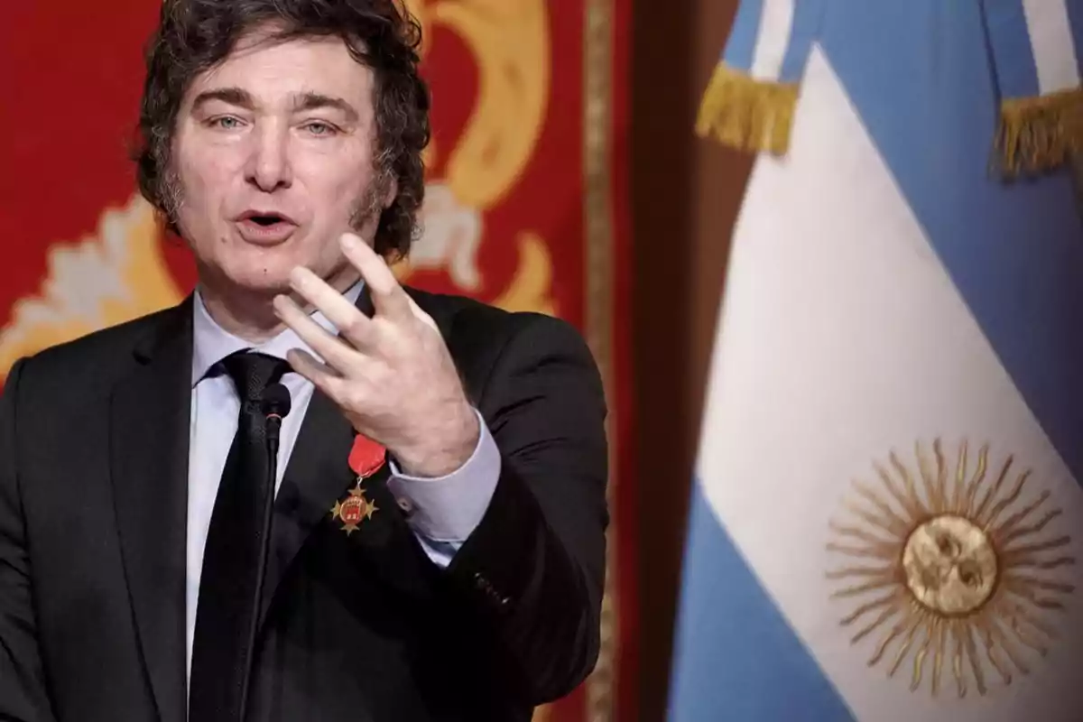 Un hombre hablando frente a un micrófono con una medalla en su traje y una bandera de Argentina al fondo.