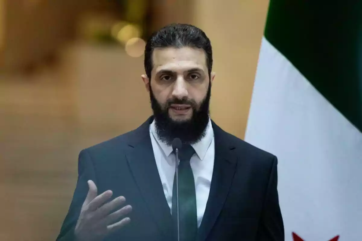 Un hombre con barba y traje oscuro habla frente a un micrófono con una bandera en el fondo.