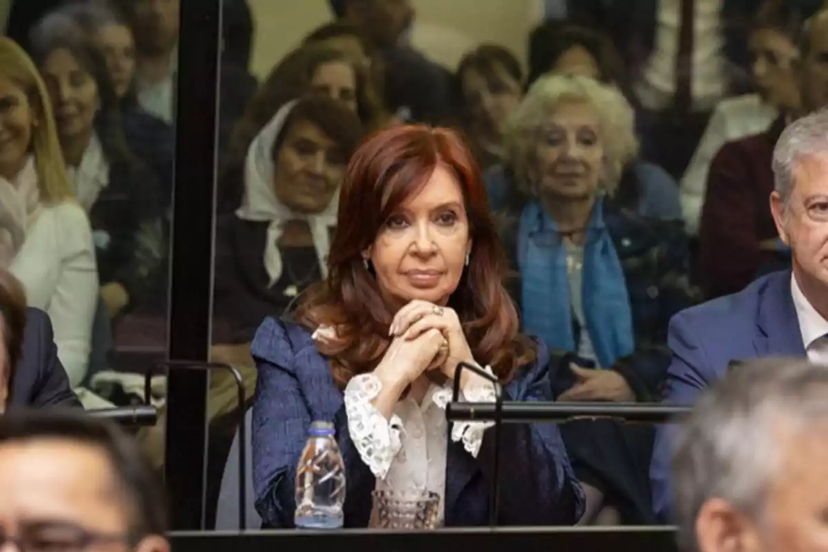 Una mujer con cabello castaño y chaqueta azul está sentada con las manos entrelazadas frente a ella en un entorno formal, rodeada de otras personas.