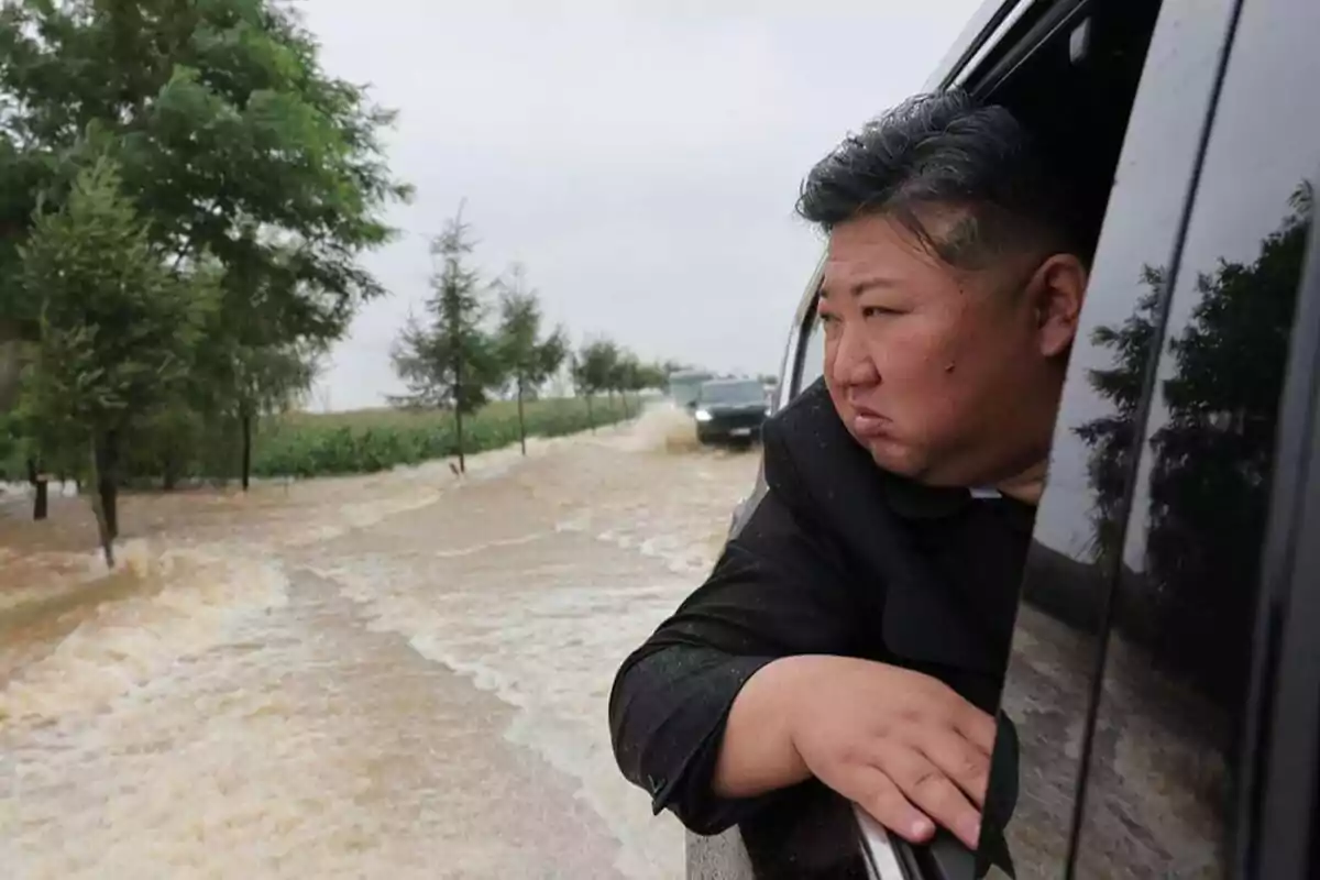 Una persona asomada por la ventana de un coche observando una carretera inundada con árboles a los lados.