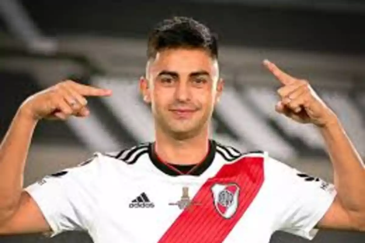 Hombre con camiseta de fútbol blanca con una franja roja y escudo, señalando con ambos dedos índices hacia su cabeza.