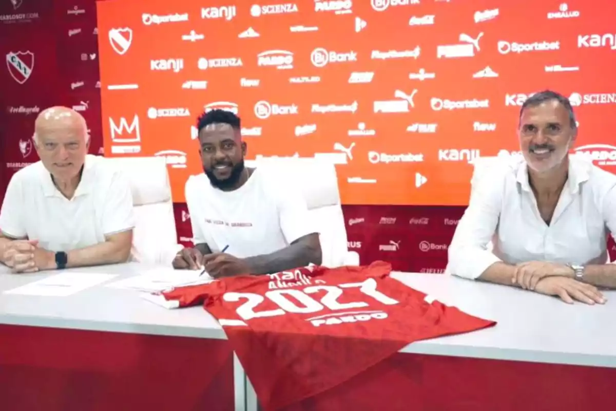 Tres hombres sentados en una mesa con una camiseta roja que dice 2027 sobre la mesa y un fondo rojo con logotipos.