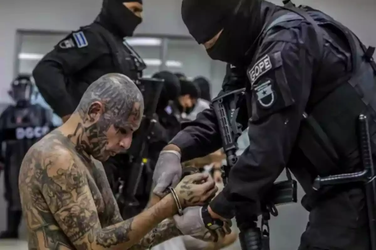 Un oficial de policía enmascarado coloca esposas a un hombre tatuado mientras otros oficiales observan en el fondo.