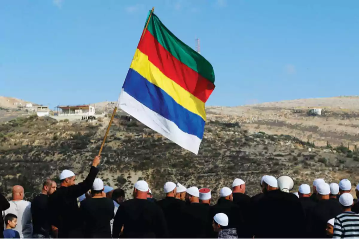 Un grupo de personas con gorros blancos sostiene una bandera de colores en un paisaje montañoso.