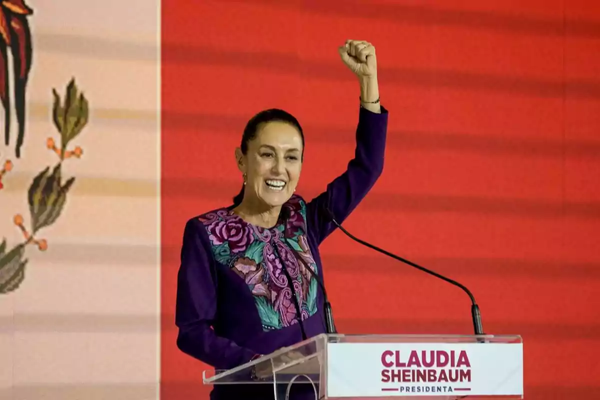 Una mujer sonriente levanta el puño mientras habla en un podio con fondo rojo.