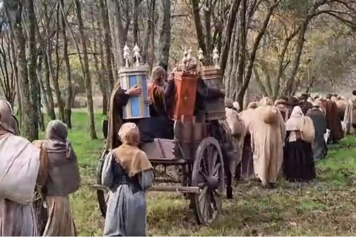 Un grupo de personas vestidas con ropas antiguas camina por un bosque llevando rollos grandes y decorados en un carro de madera.