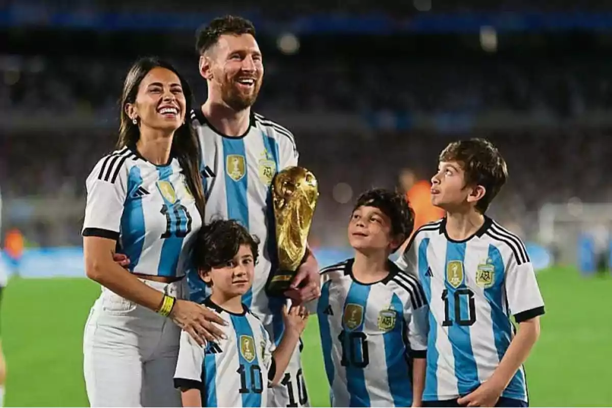 Una familia sonriente con camisetas de la selección argentina de fútbol, sosteniendo un trofeo dorado en un estadio.