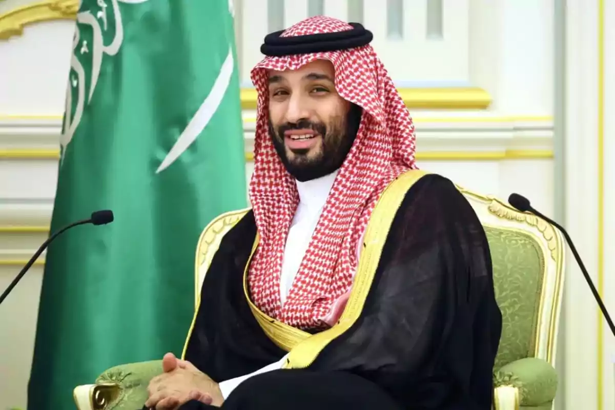 Un hombre con vestimenta tradicional saudí sentado en una silla elegante con una bandera verde de fondo.