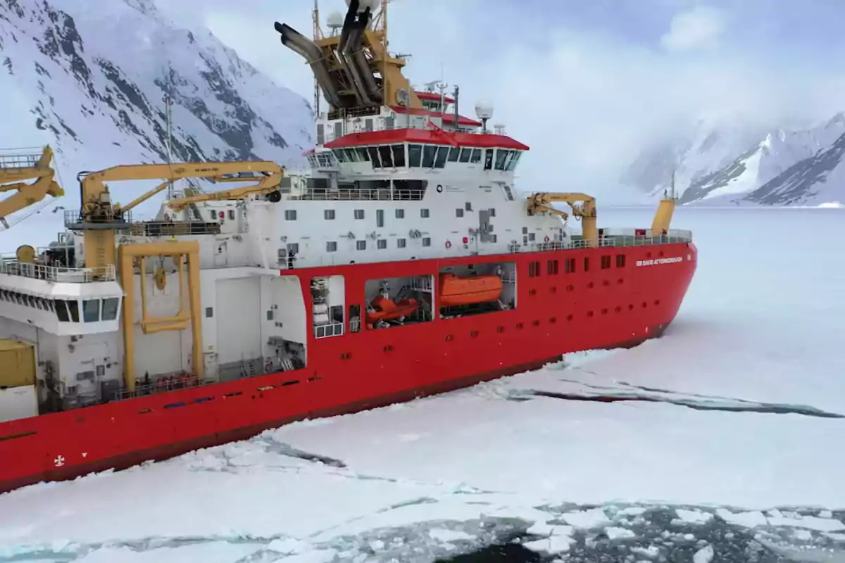 Un barco rompehielos rojo navega a través de un paisaje helado rodeado de montañas nevadas.