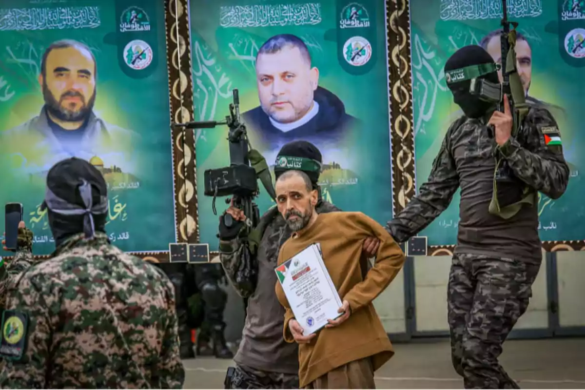 Un grupo de personas armadas con uniformes militares sostiene a un hombre que lleva un documento, mientras en el fondo se ven grandes carteles con retratos de otras personas.