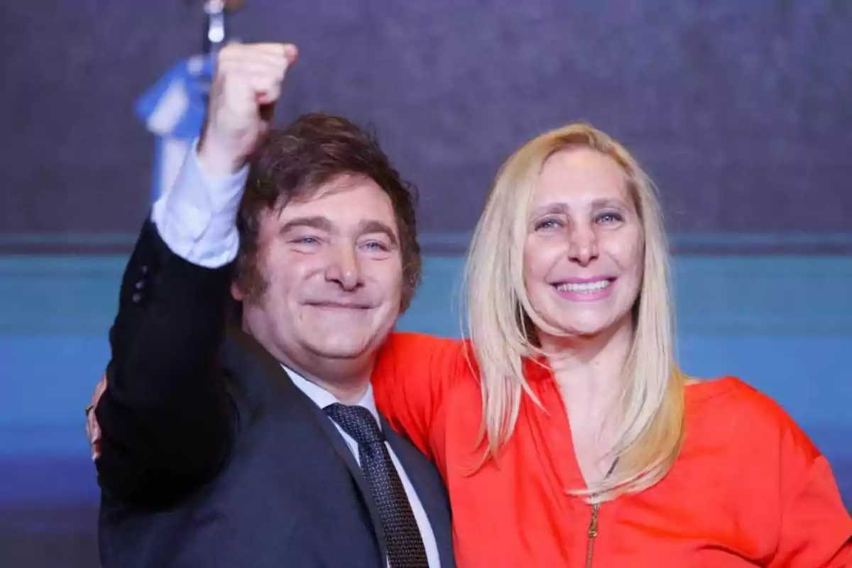 Dos personas sonrientes, una levantando el puño, posan juntas frente a un fondo oscuro con una bandera azul y blanca.