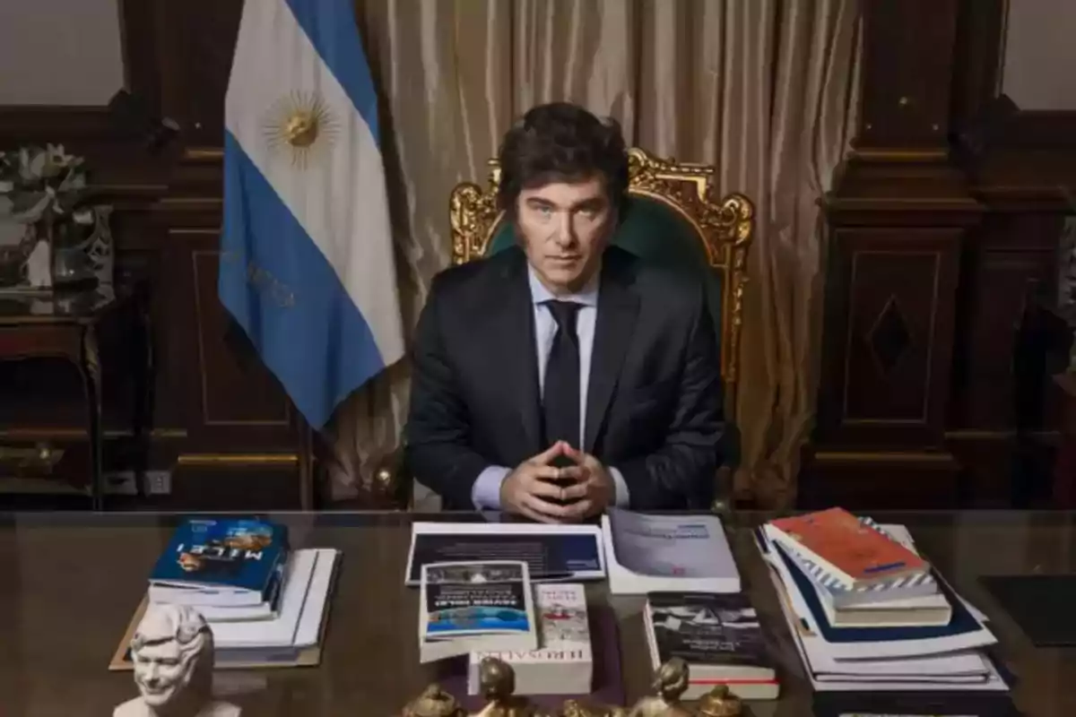 Un hombre sentado en un escritorio con libros y una bandera argentina detrás.