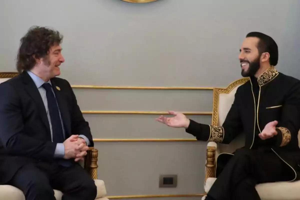 Dos hombres sentados en sillas doradas, uno con traje oscuro y el otro con traje negro con detalles dorados, conversando y sonriendo.