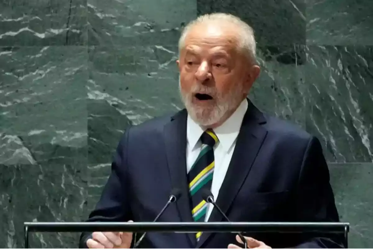 Un hombre mayor con barba y traje oscuro hablando en un podio con un fondo de mármol verde.