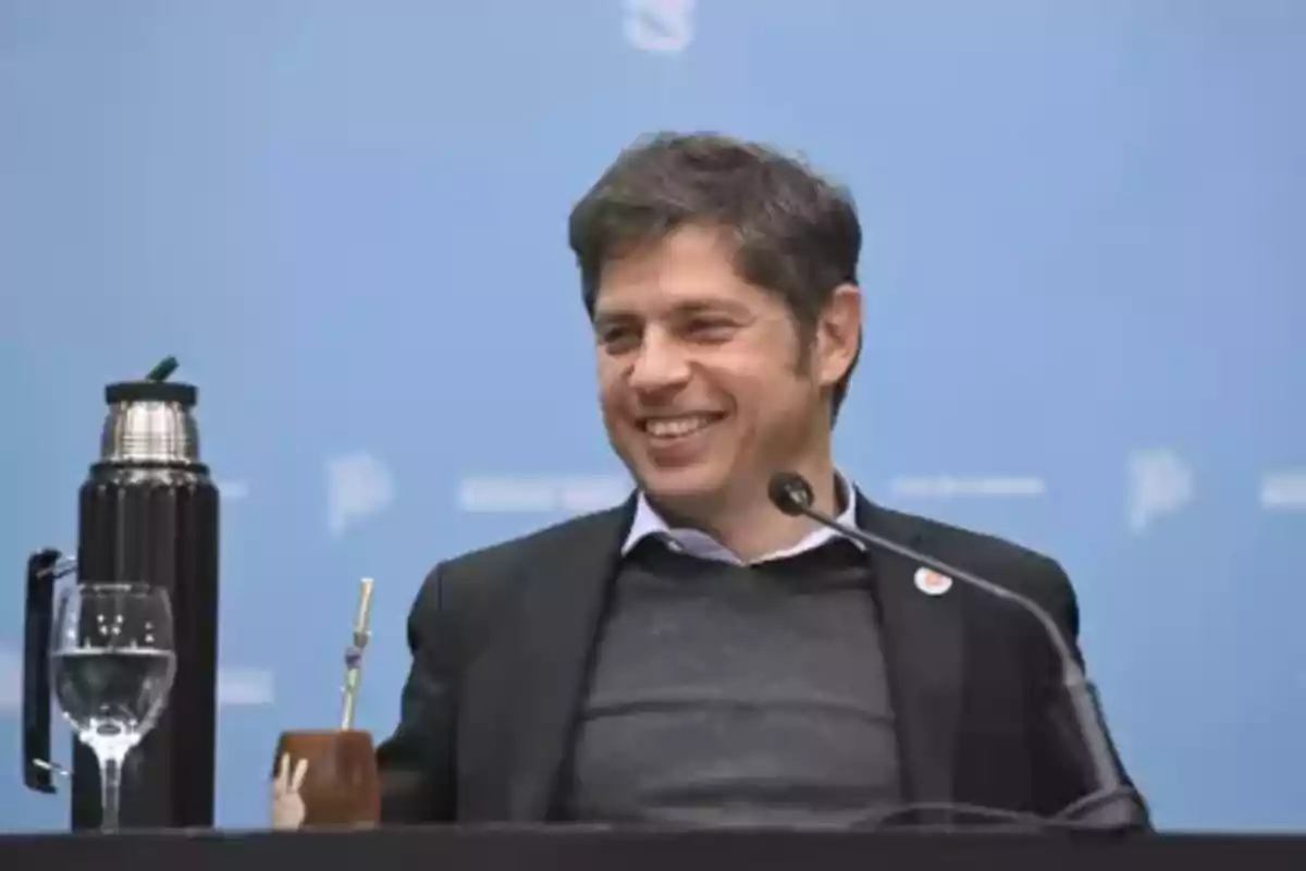 Un hombre sonriente en una conferencia con un micrófono frente a él y un termo y un mate sobre la mesa.