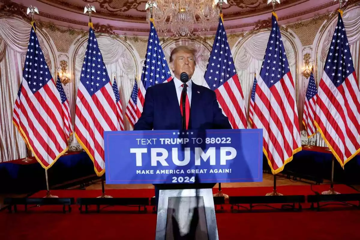 Un hombre de traje oscuro y corbata roja habla en un podio con un cartel que dice "TRUMP 2024" rodeado de banderas estadounidenses en un salón elegante.