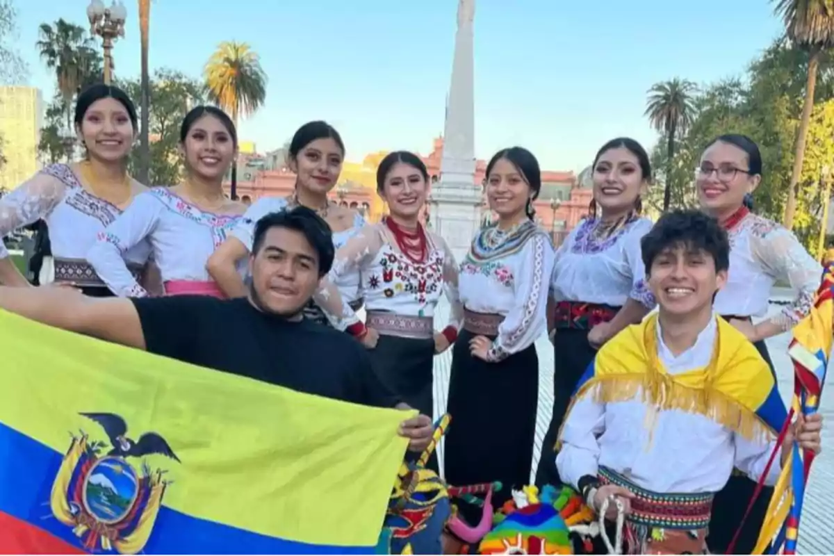 Un grupo de personas vestidas con trajes tradicionales posan con una bandera de Ecuador en un entorno al aire libre con palmeras y un monumento al fondo.