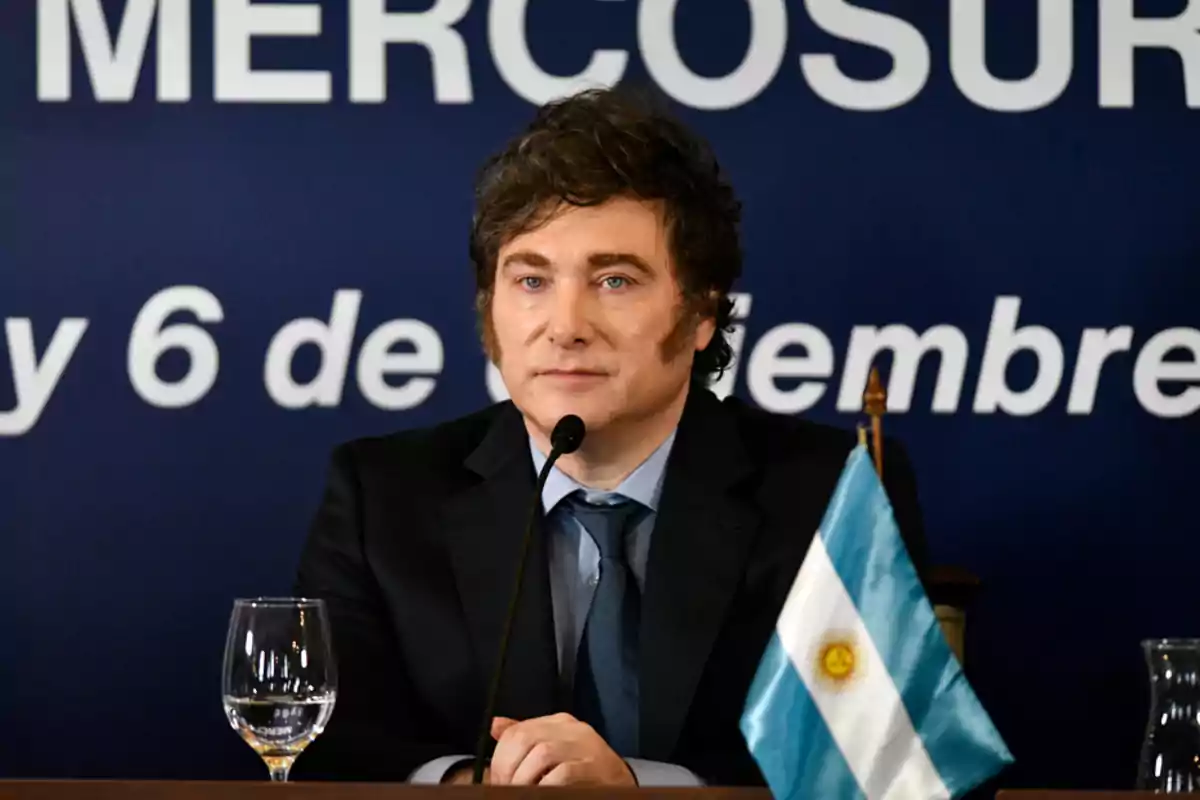 Un hombre en traje oscuro está sentado frente a un micrófono con una bandera argentina a su lado y un fondo azul con texto.