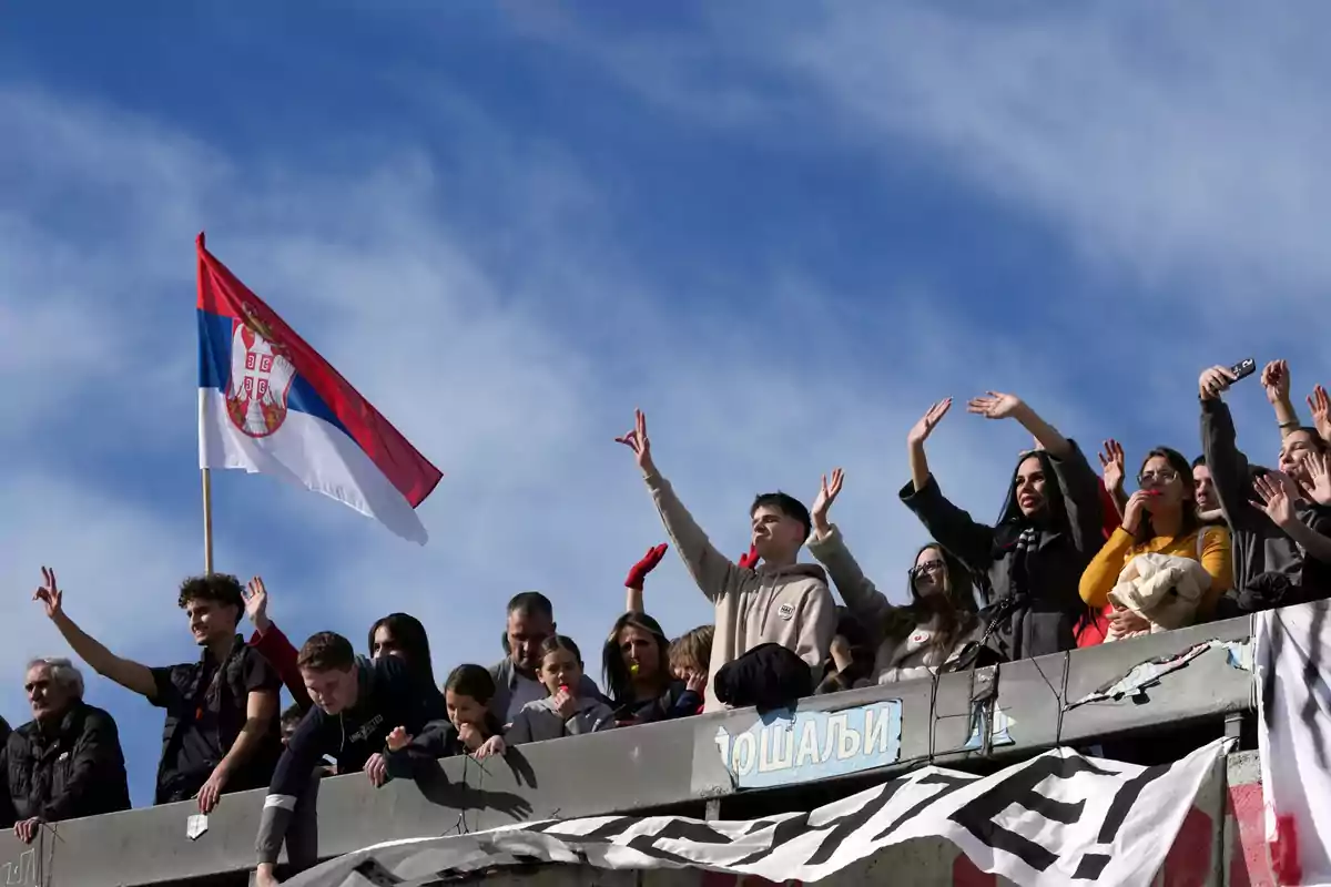 Un grupo de personas en un balcón levantando las manos junto a una bandera de Serbia ondeando bajo un cielo azul.