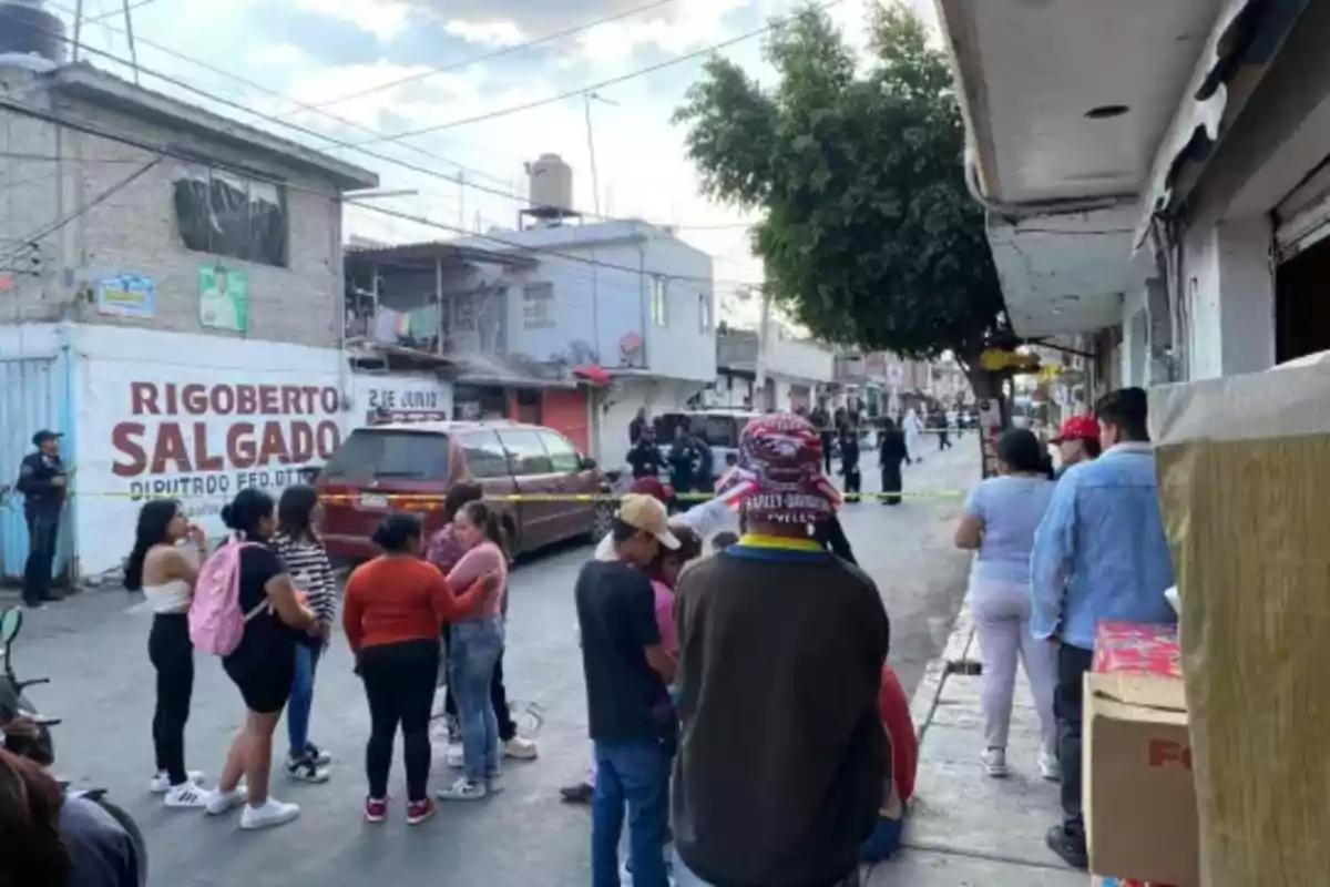 A group of people gathers on a street where there is security tape and a police presence in the background.