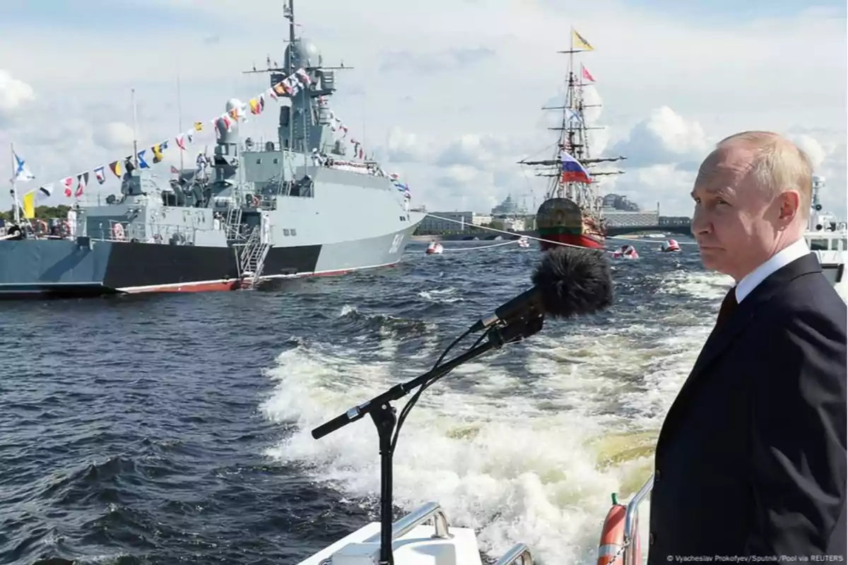 Un hombre de traje está de pie en un barco con un micrófono frente a él, mientras observa un desfile naval con barcos decorados con banderas en el agua.