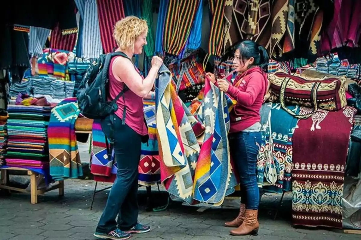 Dos mujeres conversan mientras sostienen coloridas mantas en un mercado al aire libre lleno de textiles vibrantes.