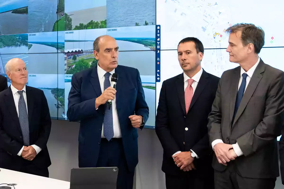 Un grupo de hombres en trajes formales está de pie frente a una pantalla grande con imágenes de paisajes y mapas, uno de ellos sostiene un micrófono mientras habla.