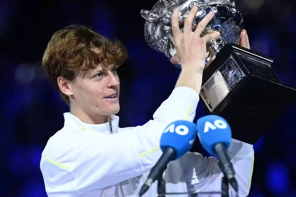 Persona sosteniendo un trofeo plateado frente a micrófonos con fondo oscuro.