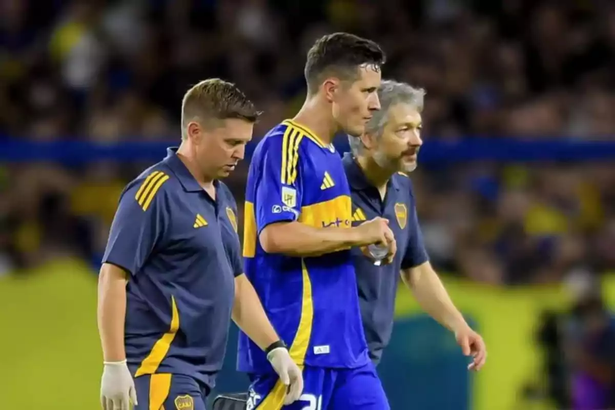 Un jugador de fútbol con uniforme azul y amarillo camina acompañado por dos personas con ropa deportiva en un estadio.