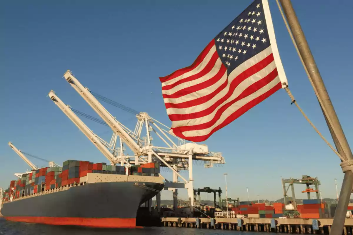 Un barco de carga con contenedores en un puerto con grúas y una bandera de Estados Unidos ondeando en primer plano.