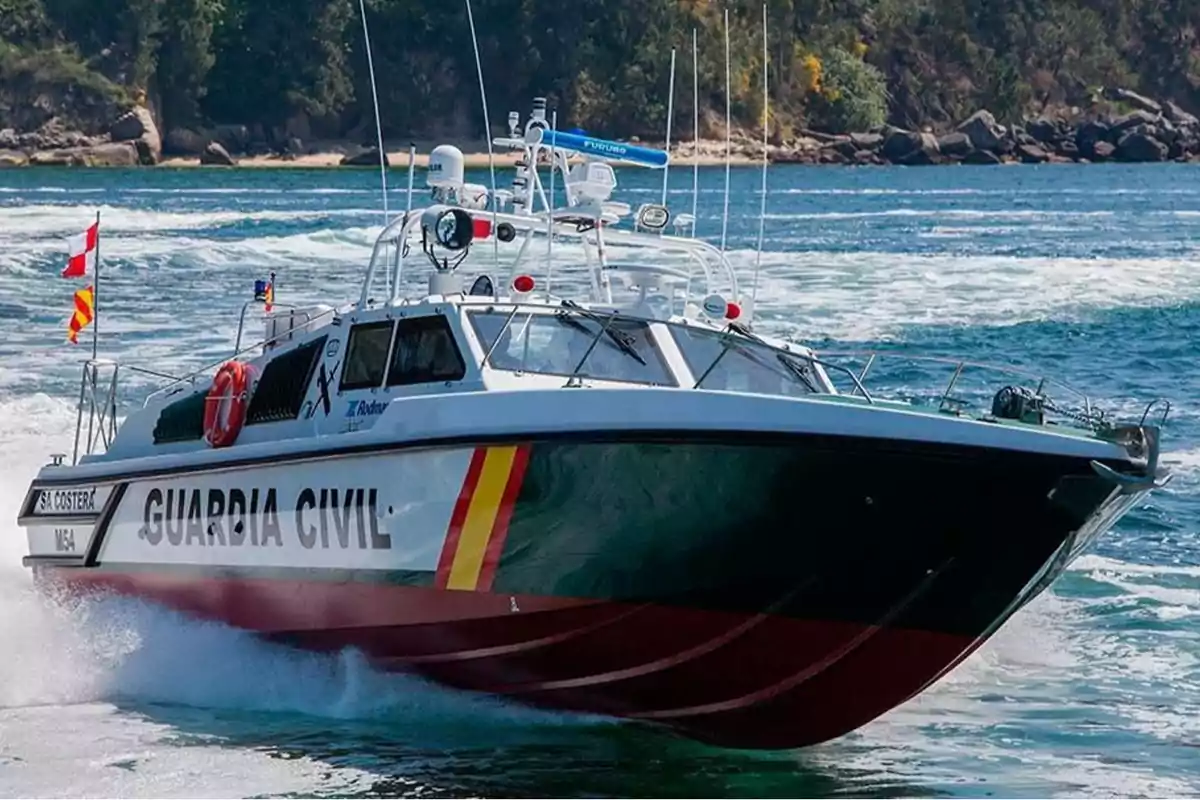 Una lancha de la Guardia Civil navegando a alta velocidad en el mar con un fondo de costa arbolada.